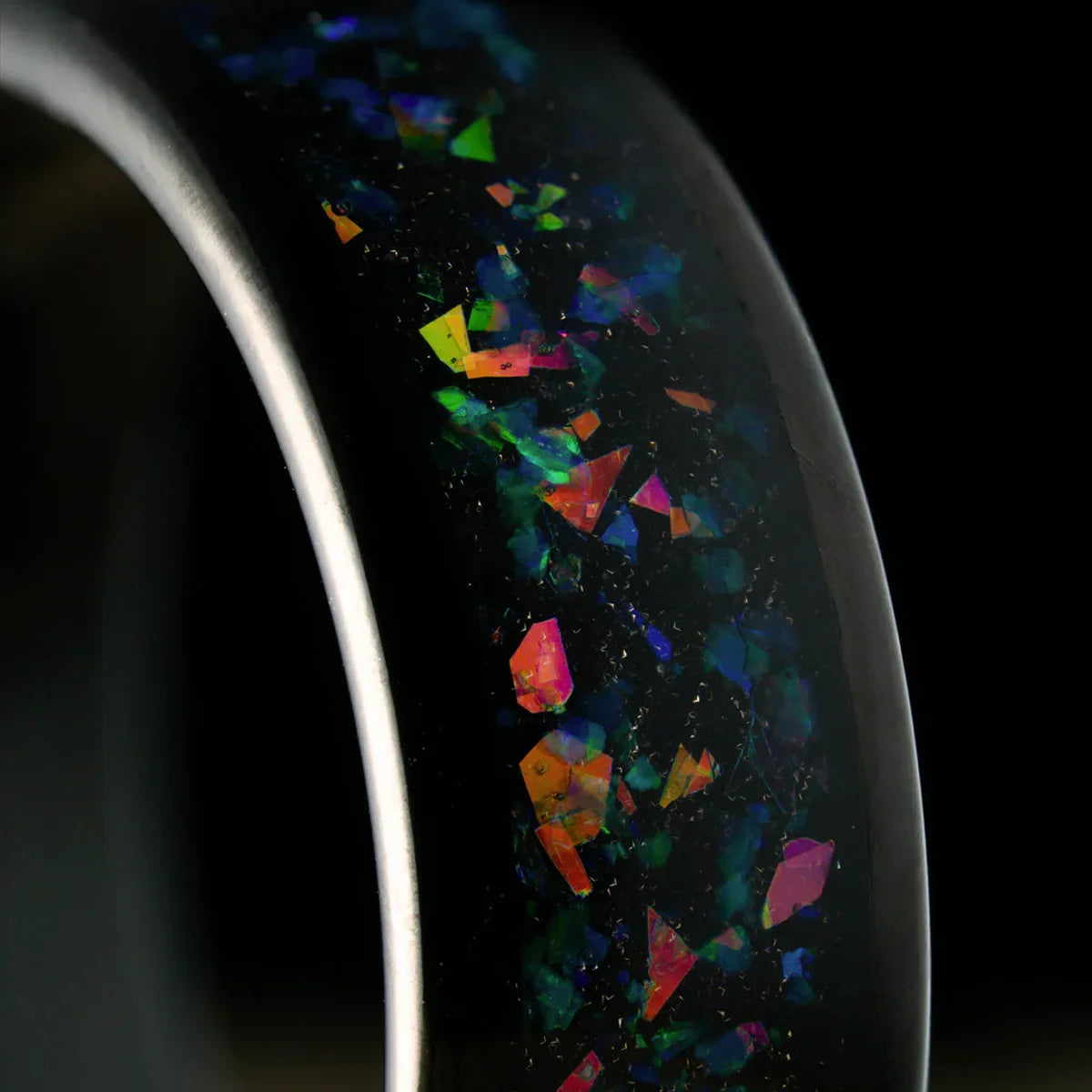 Anillo negro con incrustaciones iridiscentes multicolores, fondo oscuro.