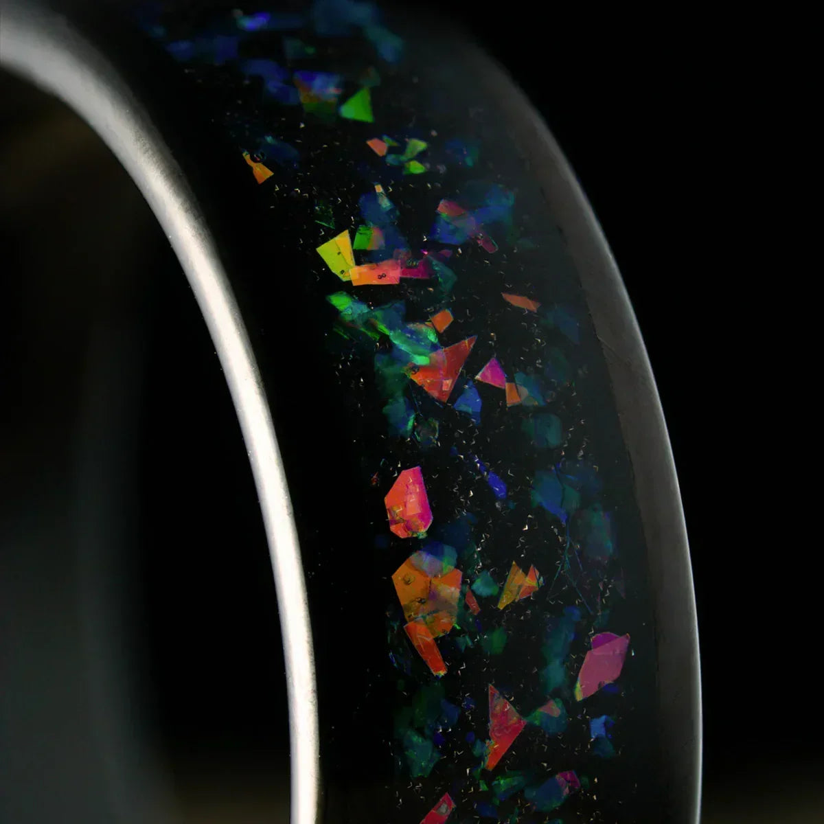 Anillo negro con incrustaciones iridiscentes multicolores, fondo oscuro.