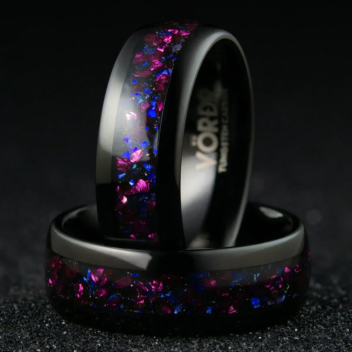 Anillo negro con incrustaciones brillantes púrpura y azul, sobre fondo oscuro, joyería moderna.