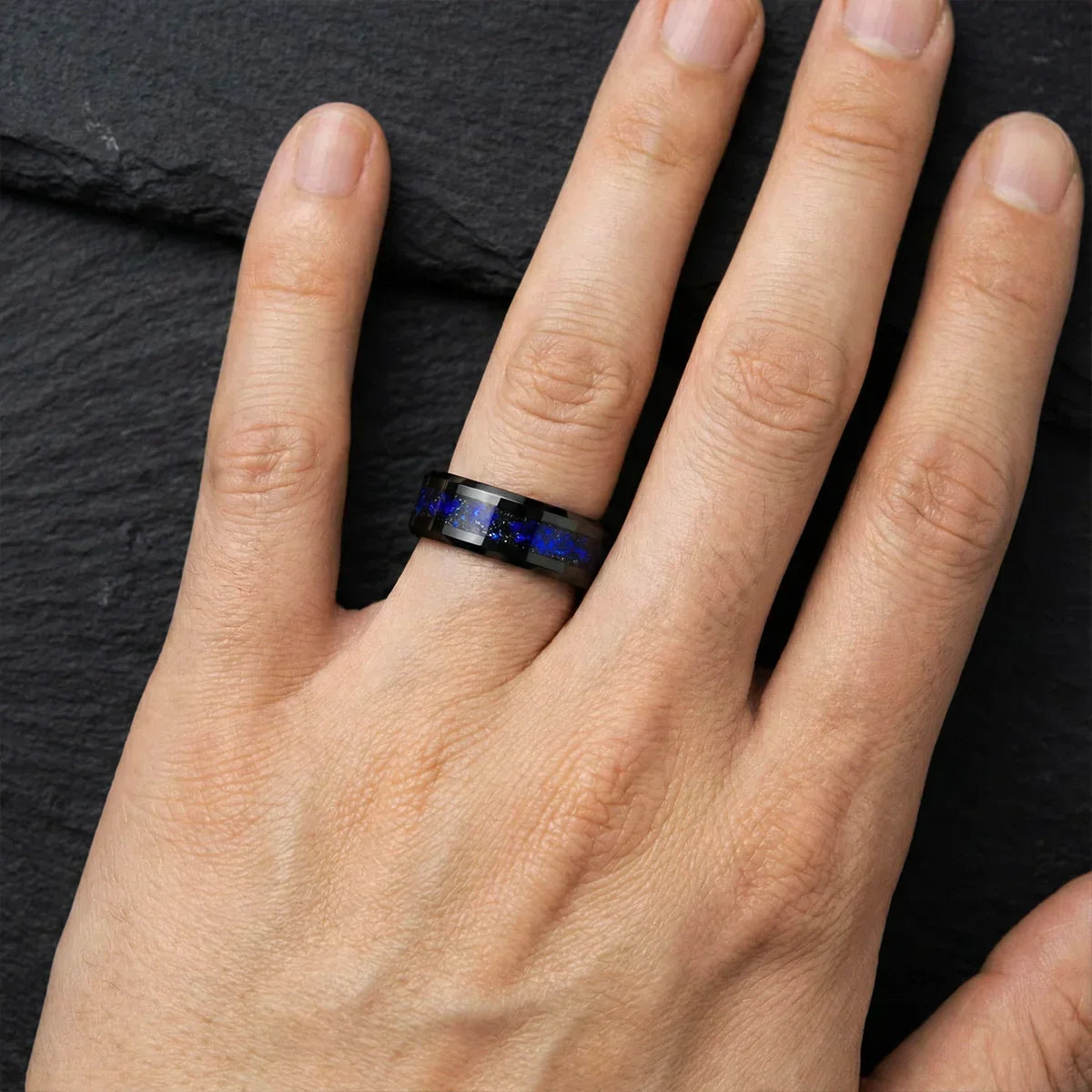 Anillo negro con incrustaciones azules brillantes en mano, fondo de pizarra.