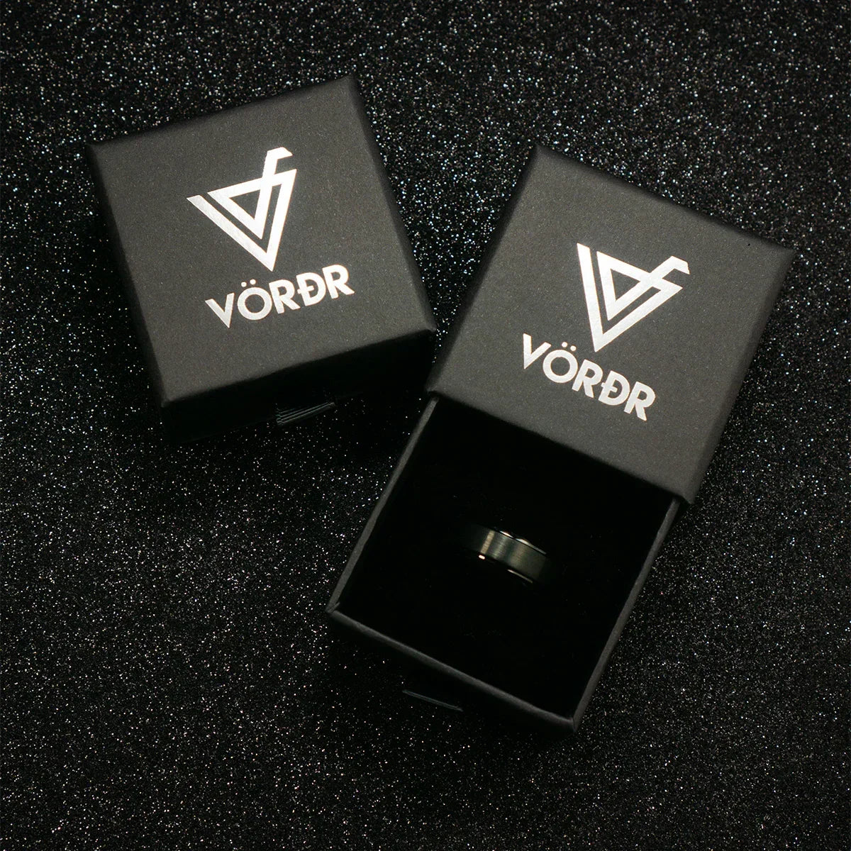 Caja negra de joyería con anillo de acero en interior sobre fondo brillante